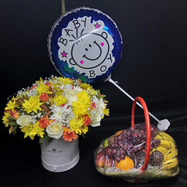 A vibrant baby boy celebration set featuring a "Baby Boy" balloon, a yellow and white floral arrangement with accents of orange flowers in a round white box, and a fresh fruit basket containing bananas, oranges, grapes, and more, wrapped in clear cellophane with a red ribbon handle.