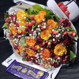 A vibrant bouquet of yellow and red roses with hypericum, baby's breath, and a Cadbury chocolate bar, beautifully wrapped in white.