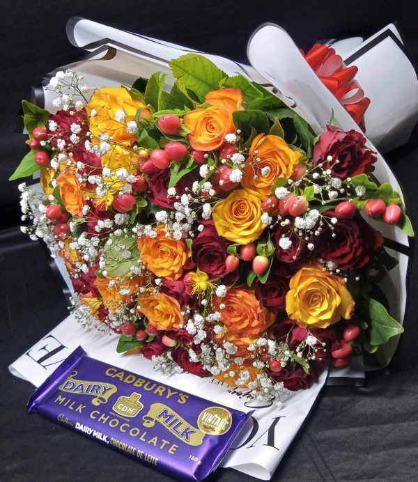 A vibrant bouquet of yellow and red roses with hypericum, baby's breath, and a Cadbury chocolate bar, beautifully wrapped in white.