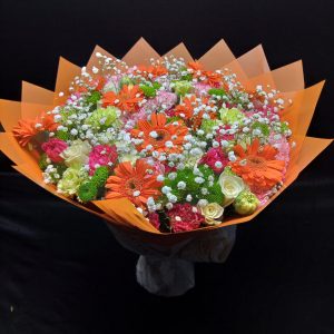 A vibrant bouquet featuring a mix of orange gerberas, green chrysanthemums, pink carnations, white roses, and baby’s breath, wrapped in orange decorative paper.